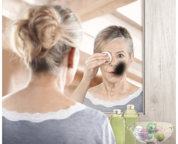 Imagen de una mujer desmaquillándose mirándose en el espejo. En el reflejo se ve una mancha negra en el rostro de la mujer, simulando lo que ve ella cuándo se mira en el espejo.